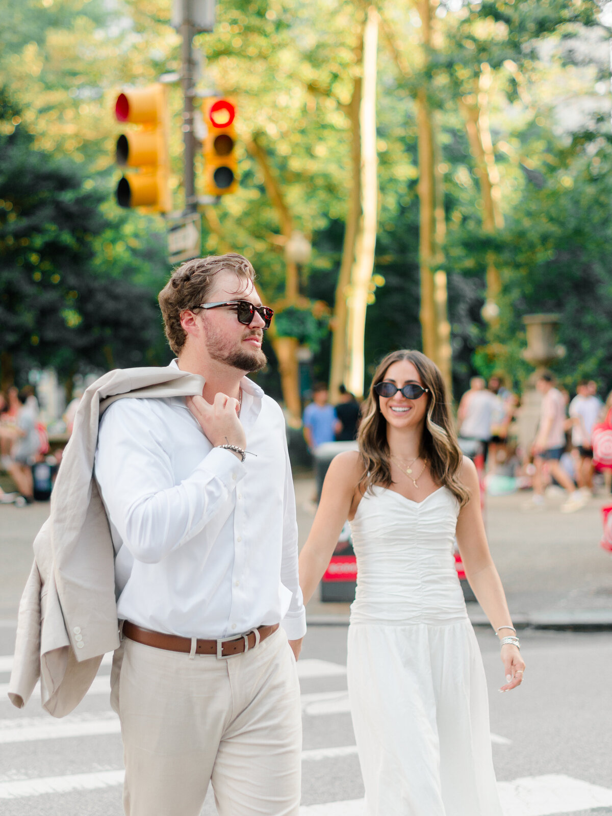 A couple walks confidently through a Philadelphia crosswalk, dressed in classic summer neutrals. The groom-to-be carries his suit jacket over his shoulder, while the bride-to-be smiles at his side in a ruched white dress and sunglasses. The golden-hour light and city bustle create a chic, modern atmosphere—captured by Elizabeth Kane Photography, a Luxury Philadelphia Wedding Photographer.