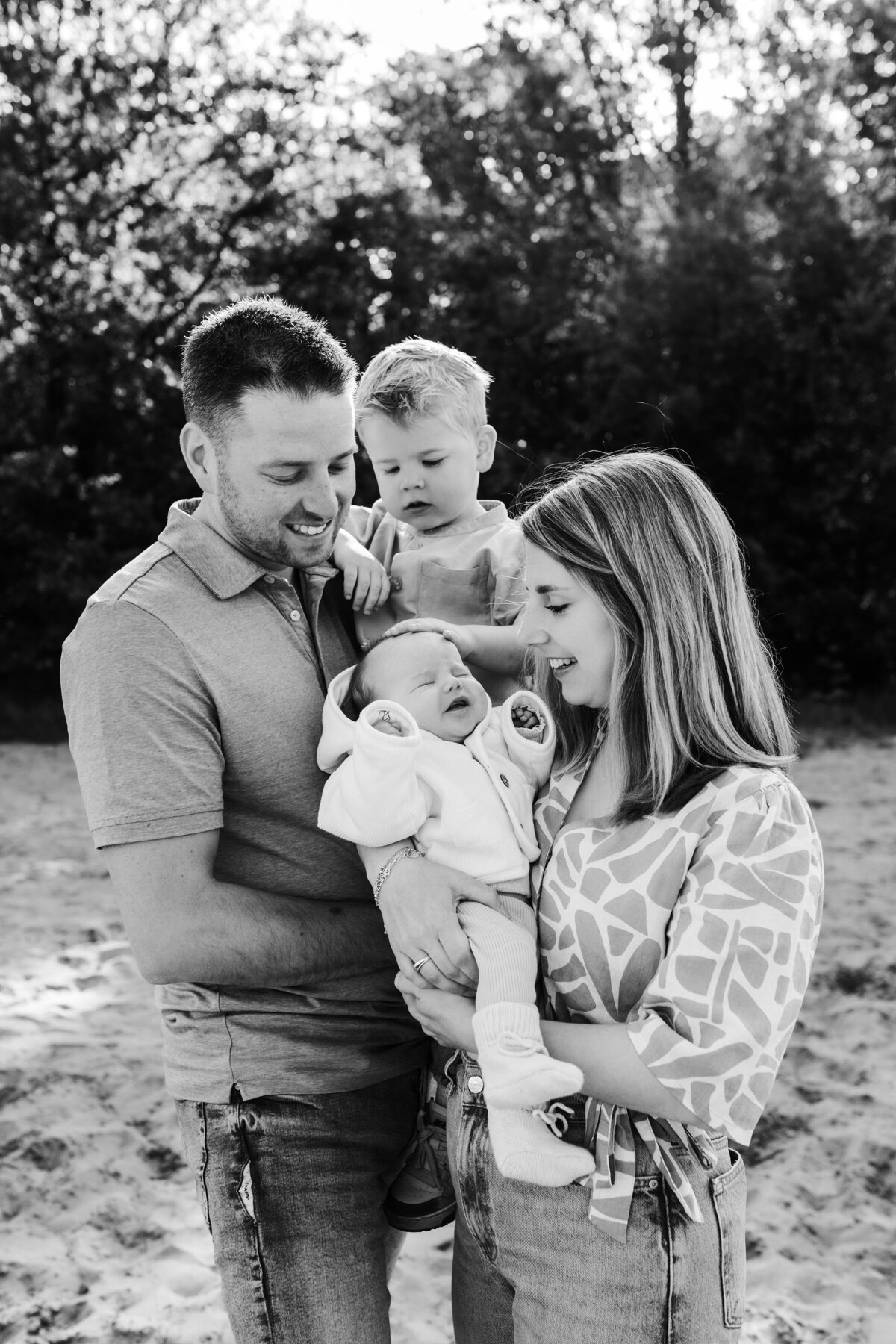 Papa houdt zoontje vast en mama baby zoontje aait over hoofd van baby en ouders kijken trots toe zwartwit