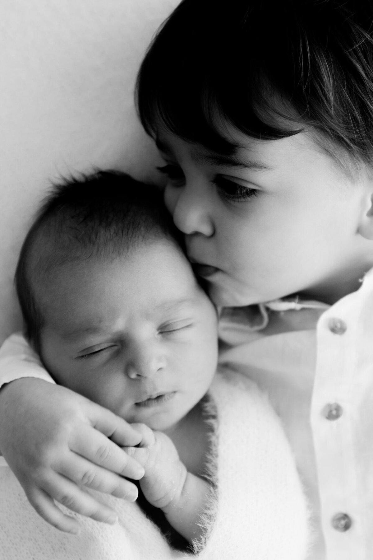 Black and white close-up of an older sibling gently kissing and cuddling their newborn baby brother or sister, captured in a soft and emotional moment.