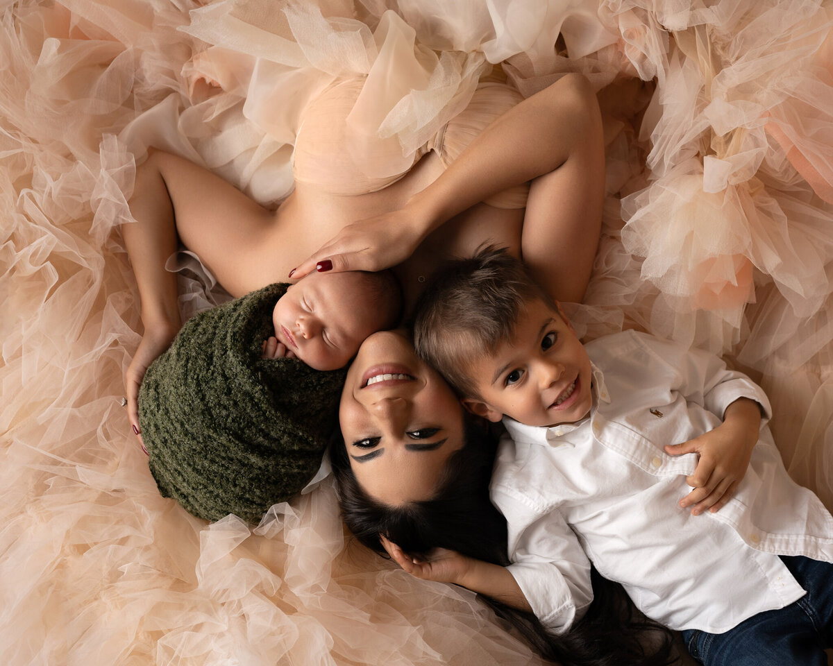 Mother lying on peach tulle gown holding newborn and smiling beside her toddler son | Elegant newborn family photography near Buda and Lakeway