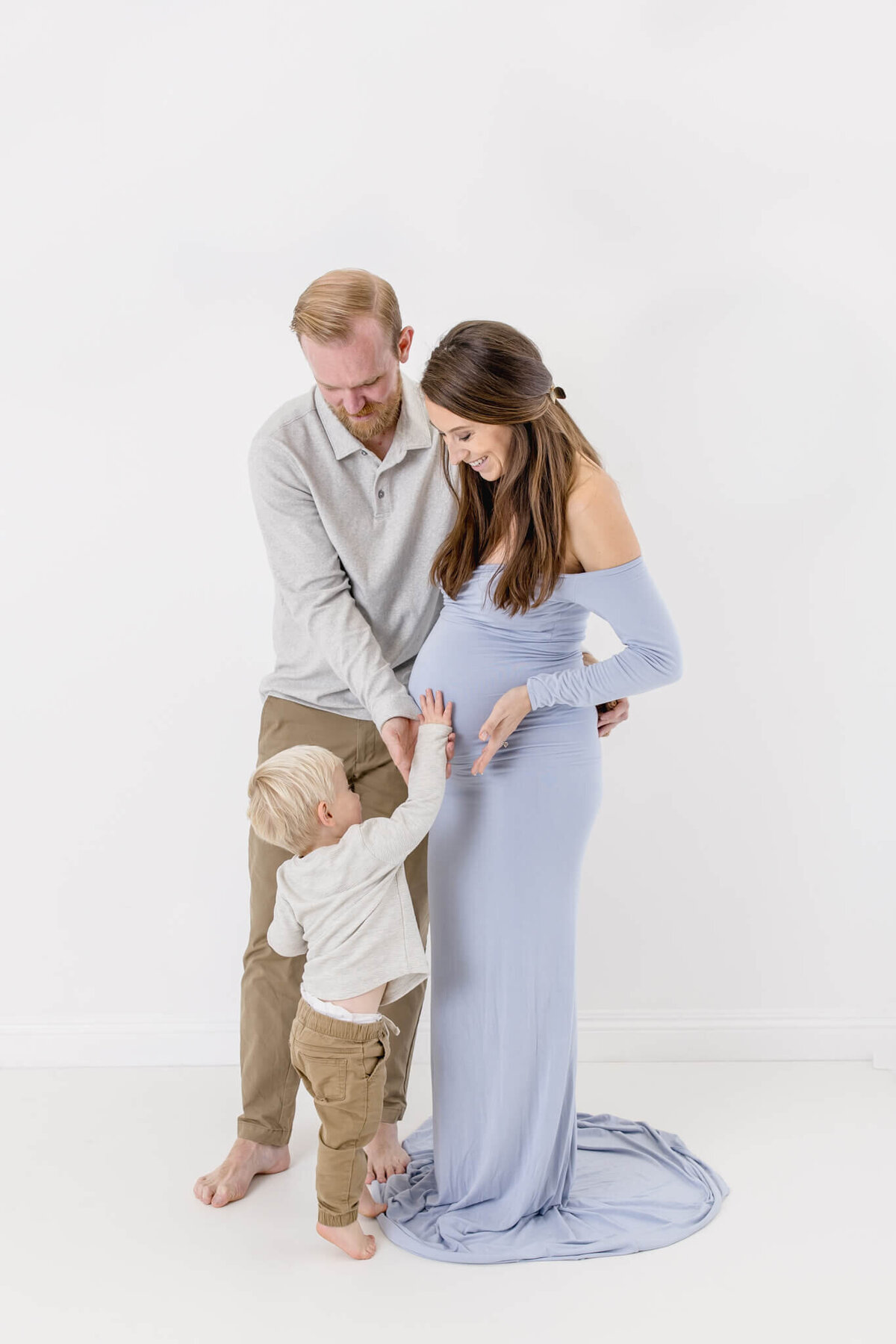 A man and a woman stand together, smiling and touching the womans pregnant belly. A young child reaches up to touch the belly too. The woman wears a light blue dress, and the man and child are dressed in casual clothing.
