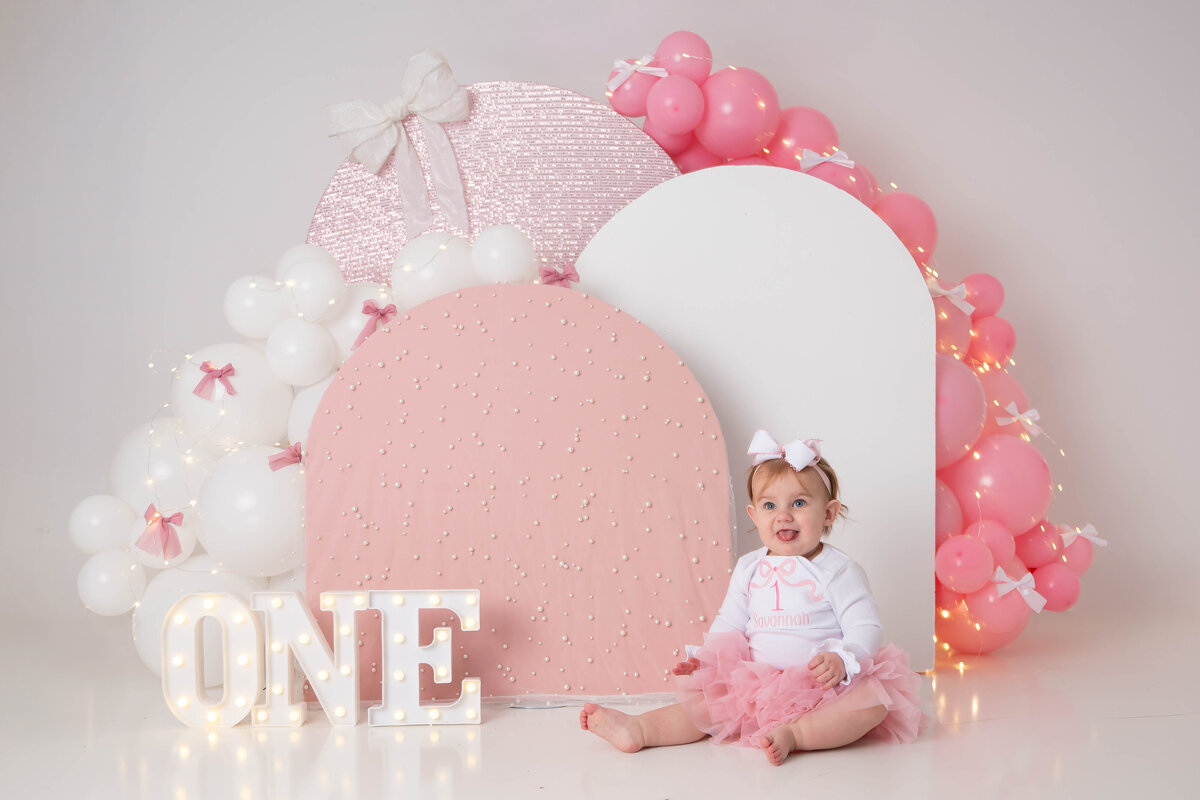 Baby in a pink tutu sits near a pink and white arch while sticking out her tongue. 