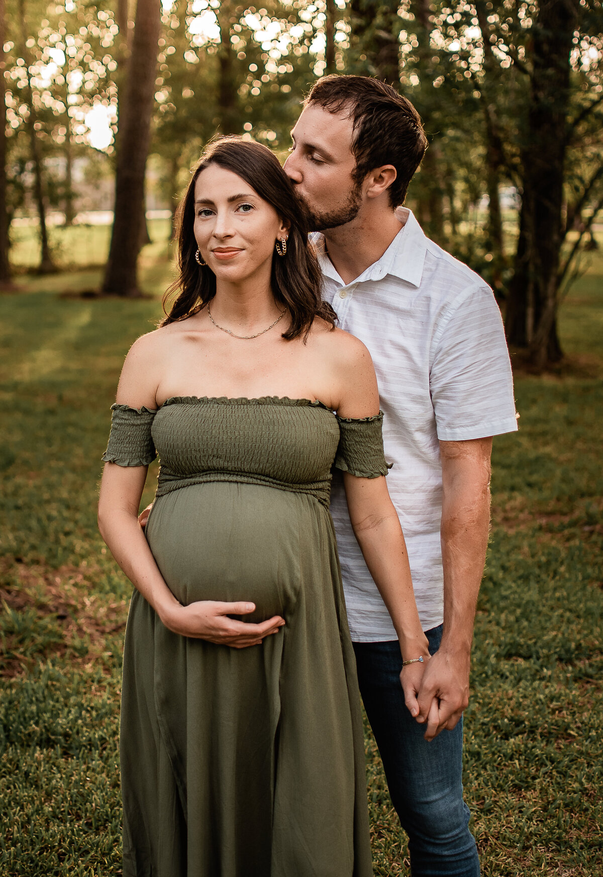 A mom to be wearing green smiles at the camera as her husband kisses the side of her head.