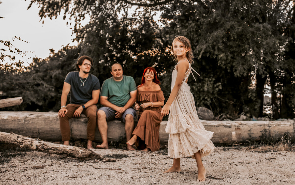 Daughter is dancing up front , parents and older brother is sitting down on the tree log , they are on the beach in Toledo