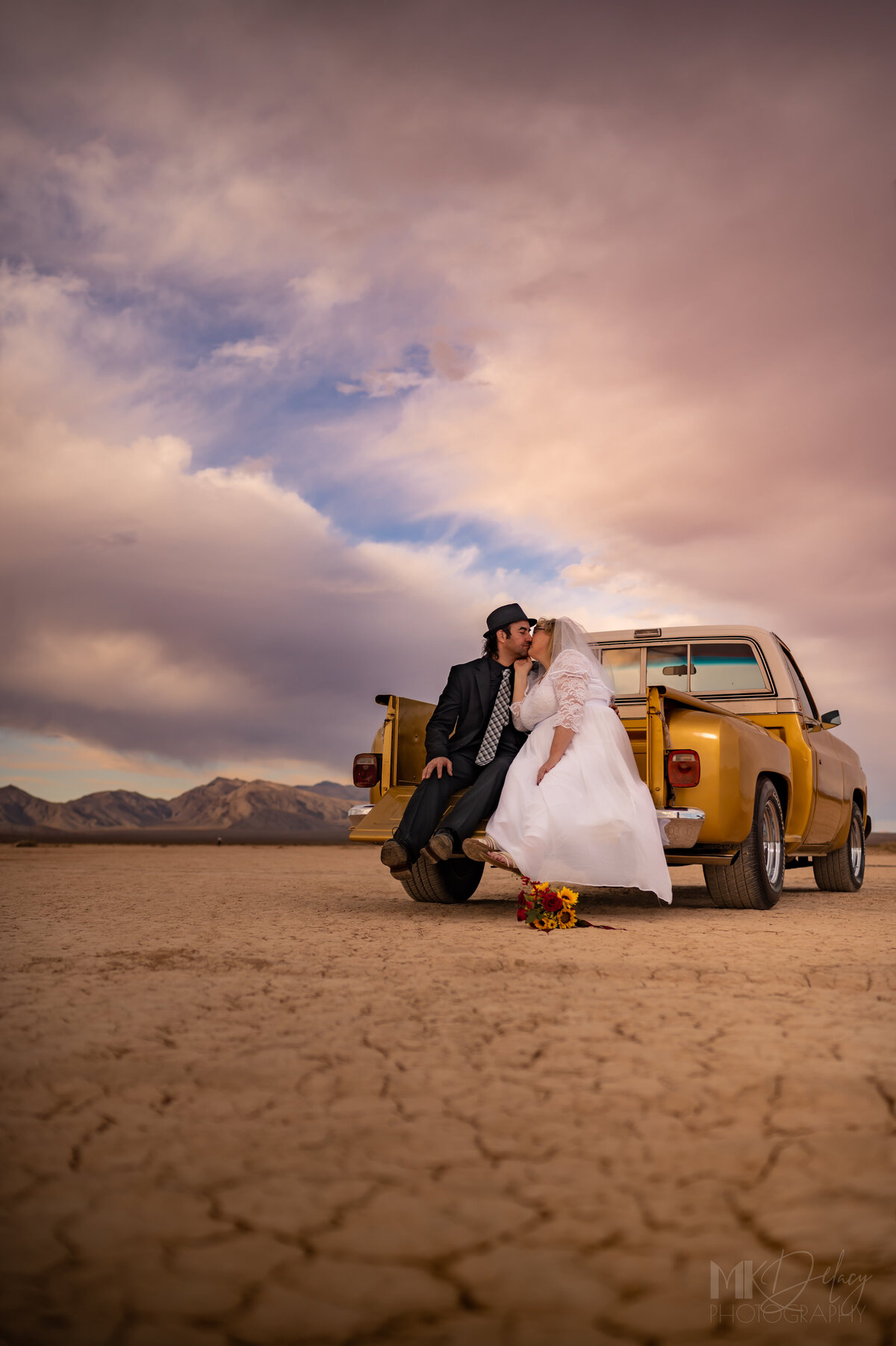 bride and groom sit on the tailgate of his gold antique remodeled checy on a dry lake bed Dry lake bed elopement bride in lace wedding gown with glasses groom in black suit white dress shirt with  fedora  classic chevy truck gold  restored with golden hour sunlight las vegas wedding photography las vegas elopement las vegas wedding photographers mk delacy photography