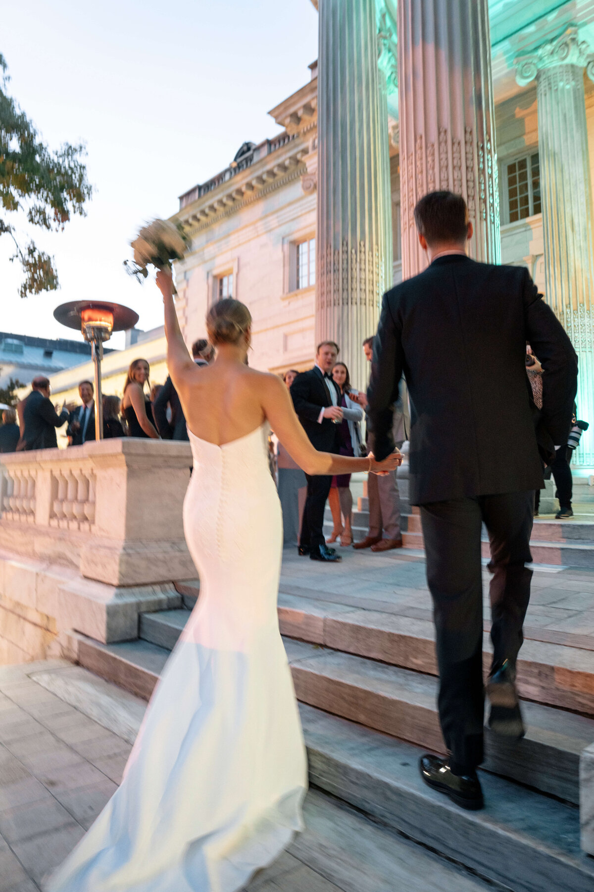 A bride and groom in formal attire ascend stairs toward a large building with columns, followed by guests. The bride holds a bouquet high in the air, and the scene is warmly lit in the evening.