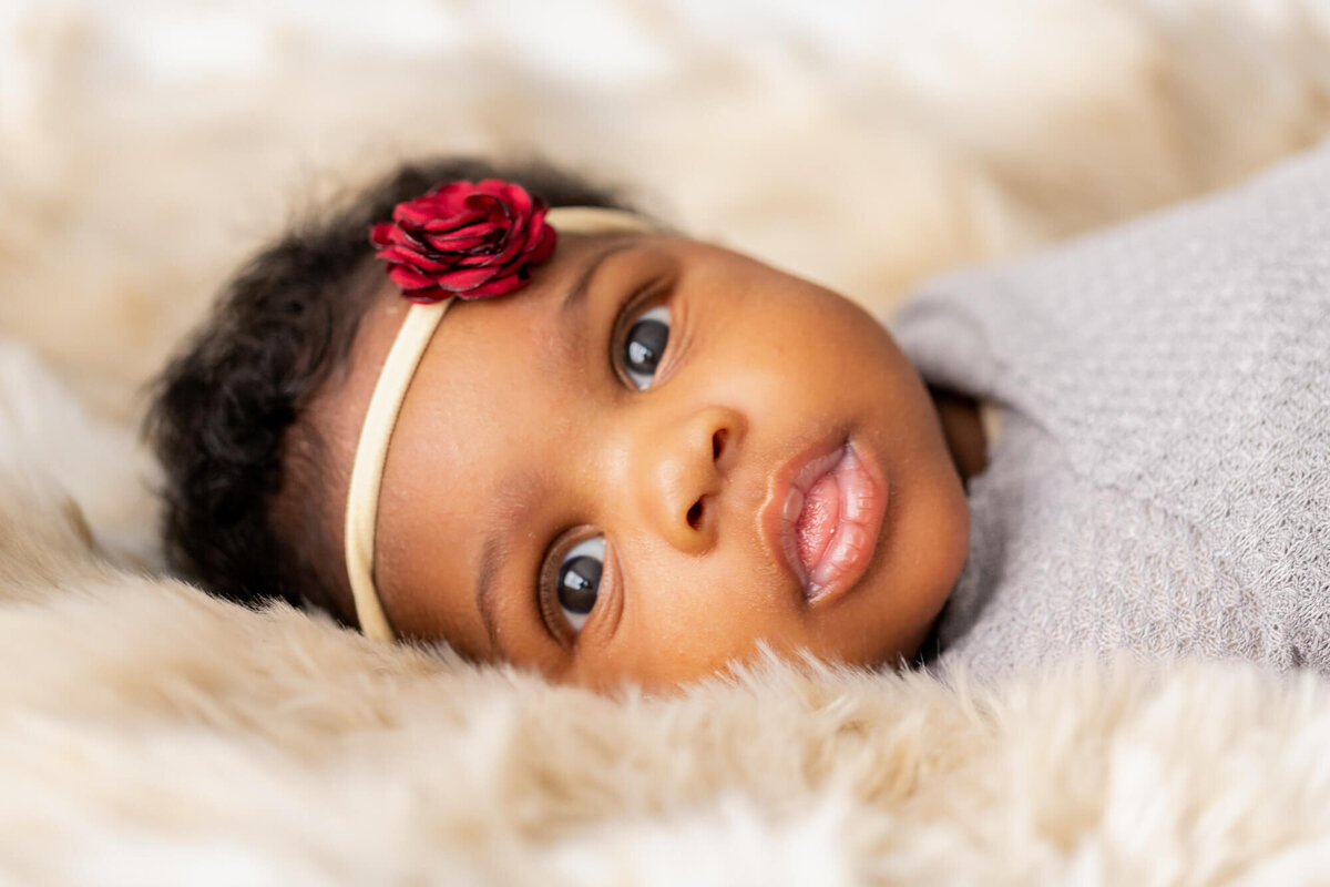 Newborn baby girl in a grey wrap and red flower bow laying on a fuzzy blanket