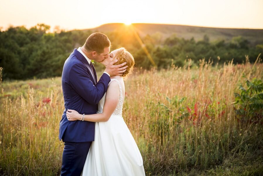 A groom holds his bride's face as the two kiss in a field at golden hour.