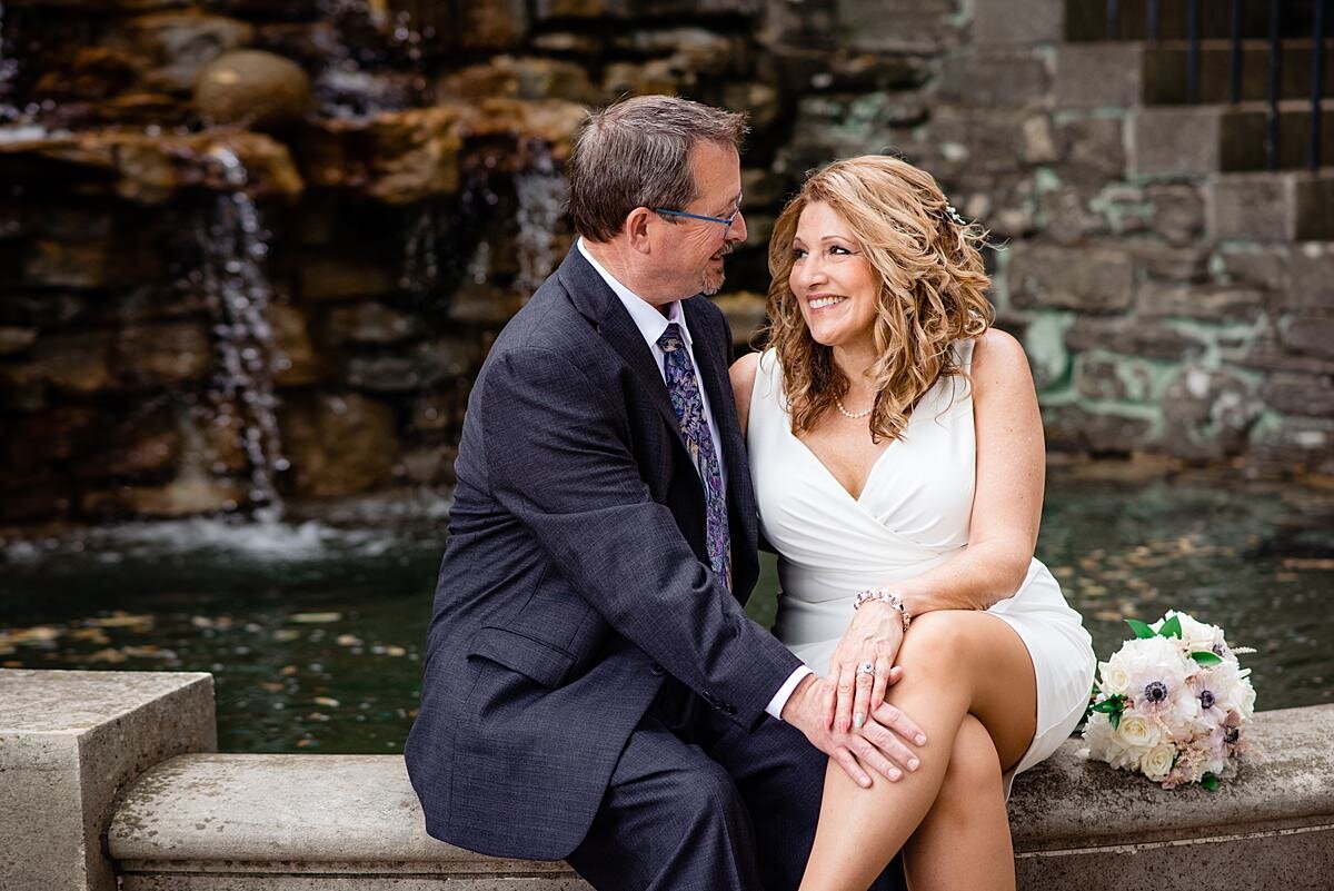 The bride and groom stare lovingly into each other's eyes as they sit at the edge of a fountain. The groom has his hand on the bride's knee and she has her hand on top of his. The groom is wearing a dark gray suit with a white shirt and blue pattered tie. The bride is wearing a short fitted white dress with a plunging neckline. Her bridal bouquet sits on the edge of the stone fountain next to her.