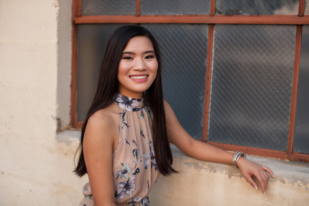 A lovely Asian American teen girl poses for a senior portrait in front of an industrial downtown window. Captured by Springfield, MO senior photographer Dynae Levingston.