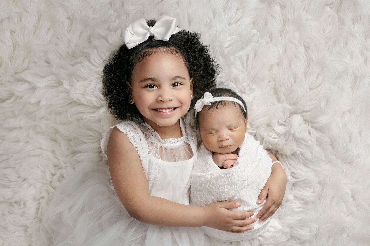 Newborn sibling photo with  a newborn baby and big sister holding him  on a white rug