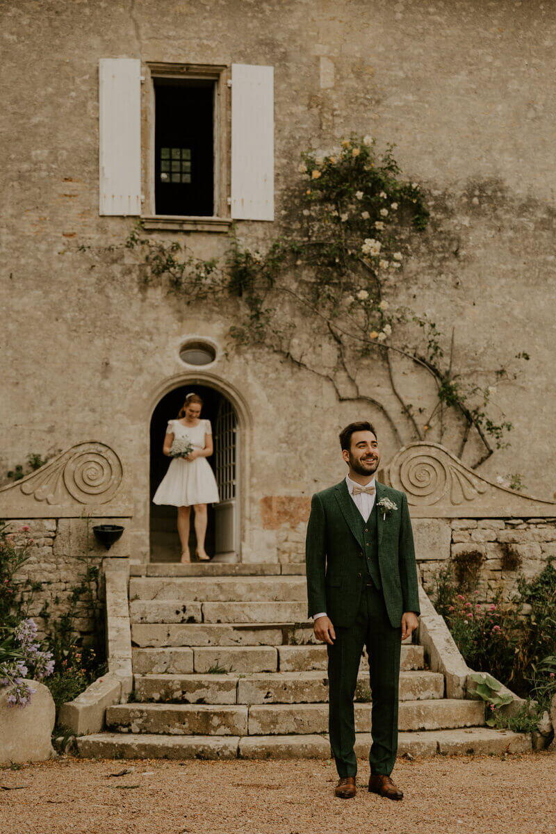 Marié attendant de découvrir la mariée sortant d'un bâtiment en pierre et s'apprêtant à descendre les marches. Photo prise par Laura Termeau, photographe professionnelle en vendée.