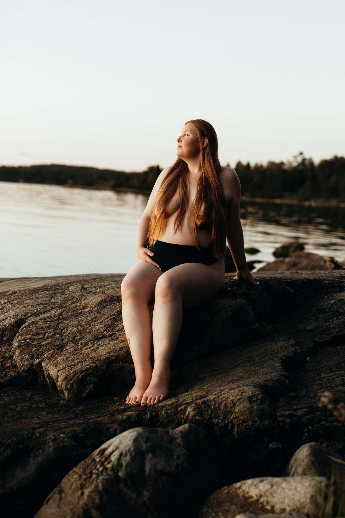 En gravid kvinna sittandes på ett berg ute vid vattnet och tittar ut mot vattnet under en gravidfotografering i Stockholm