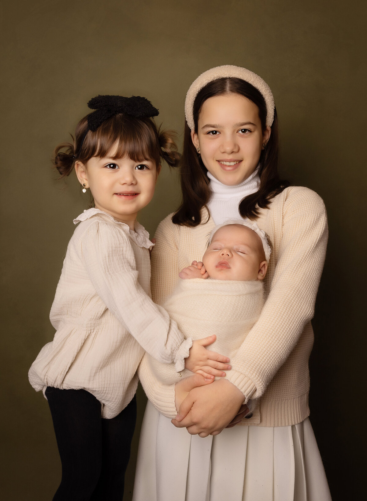 Two big sisters pose with their new baby sister for a newborn photoshoot. Big sister is holding baby and the toddler sister is standing beside her, both touching baby. The big sisters are smiling at the camera. Captured by top Brooklyn newborn photographer Chaya Bornstein