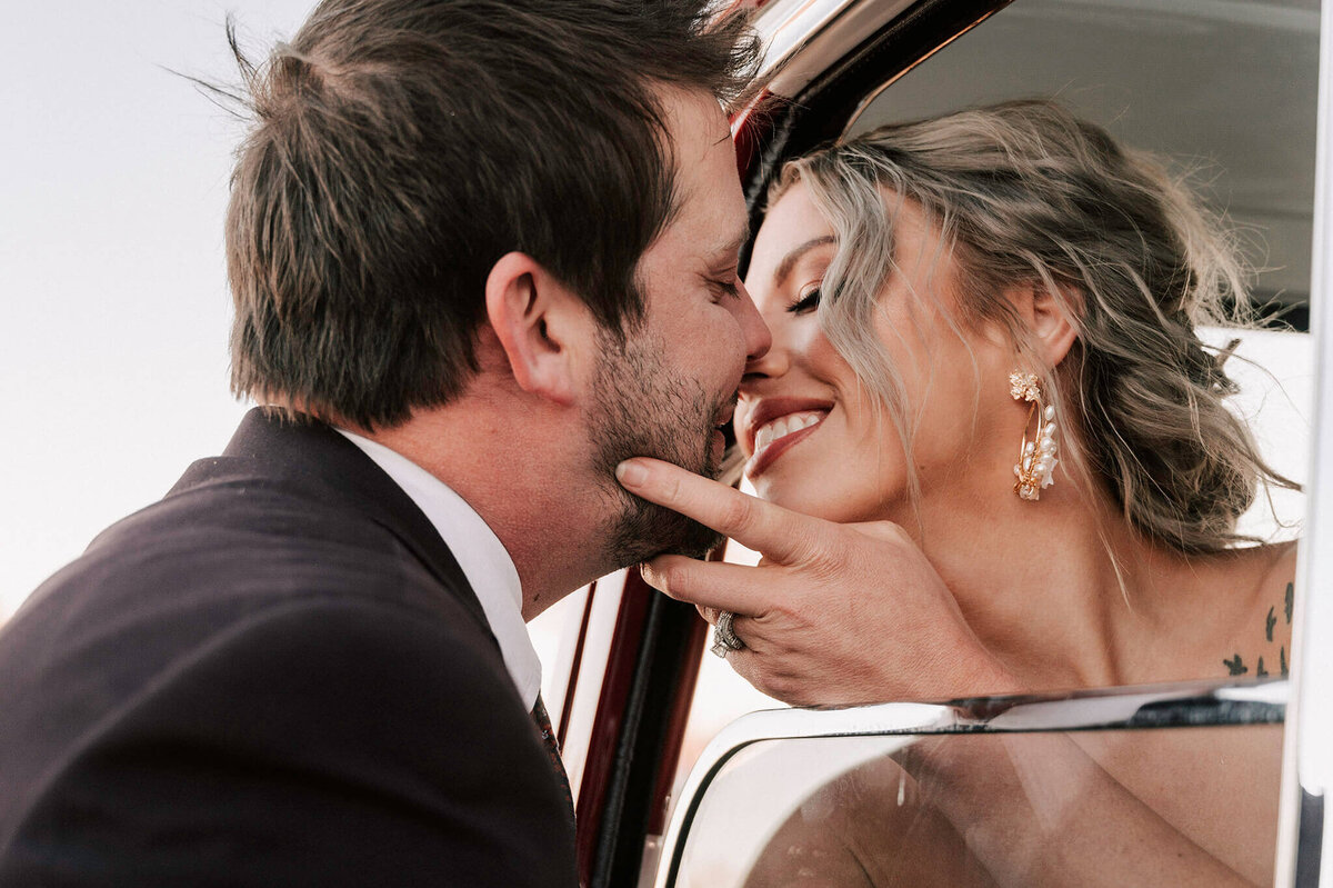 Bride hanging out of a truck window with her hand on her groom's chin who is leaning in to kiss his bride captured by meraki rose photography