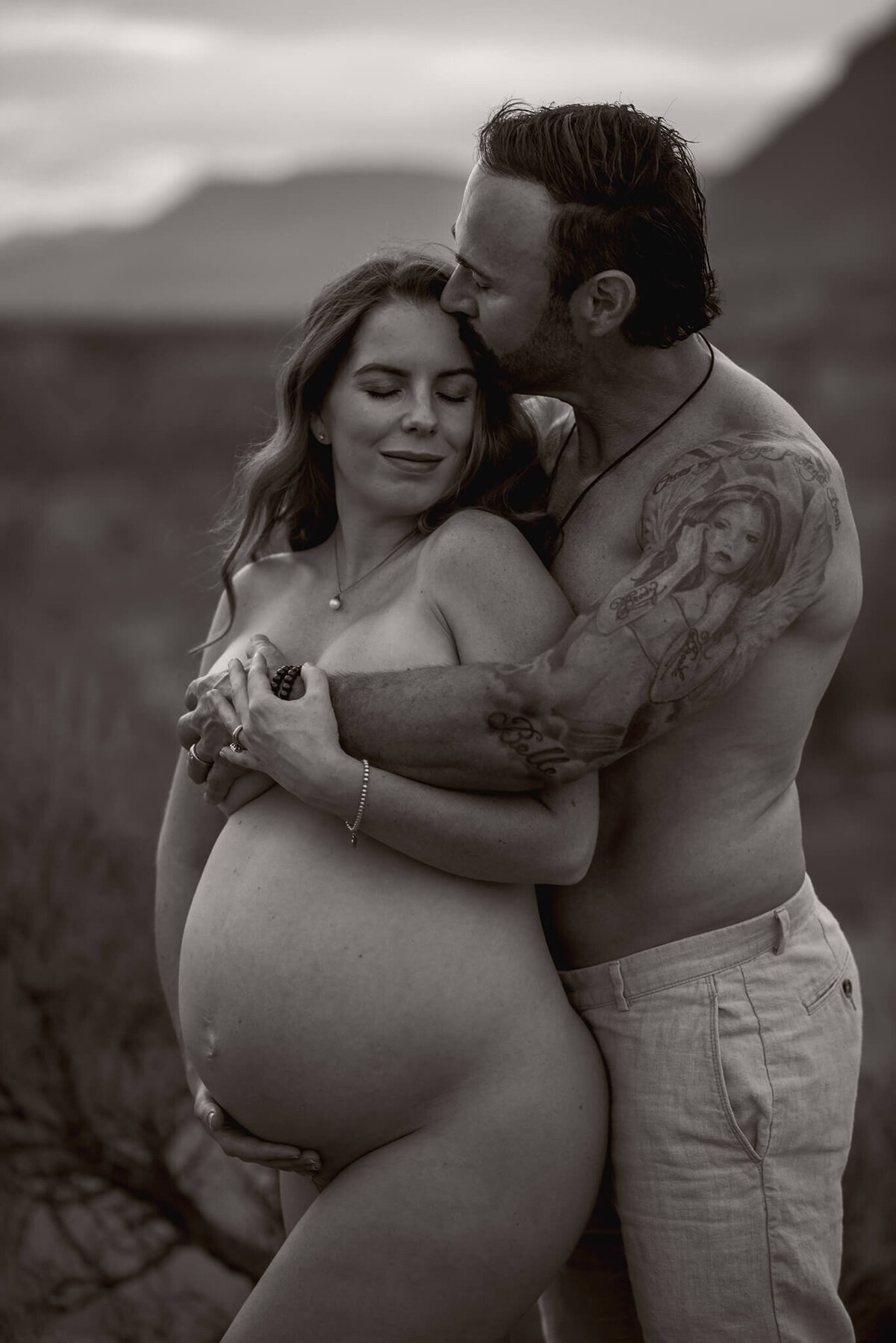 A black and white image of a shirtless man embracing his nude pregnant wife from behind. She closes her eyes lovingly while he holds her.