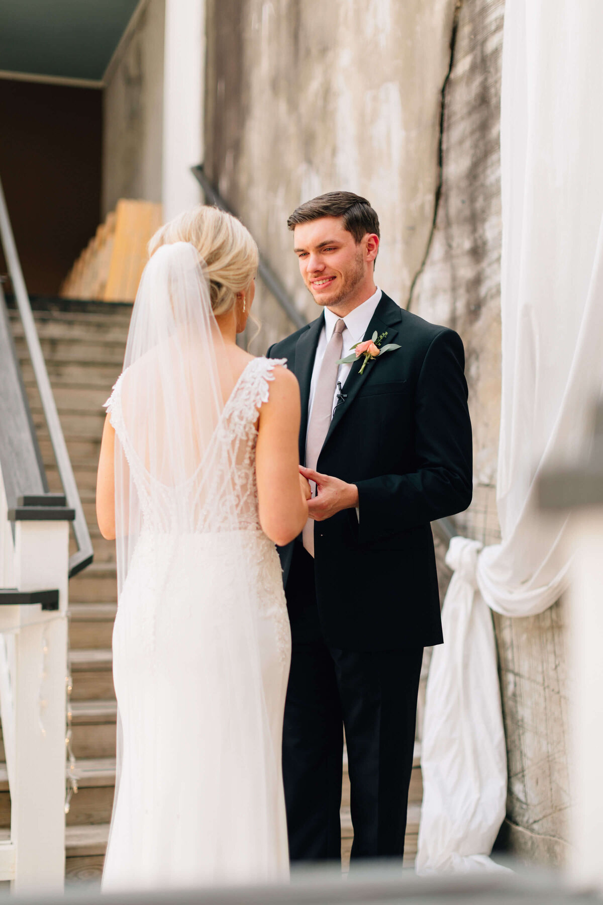 A bride in a white dress and veil smiles at a groom in a black suit and tie. They stand next to a rustic brick wall and wooden stairs, sharing a joyful moment together.