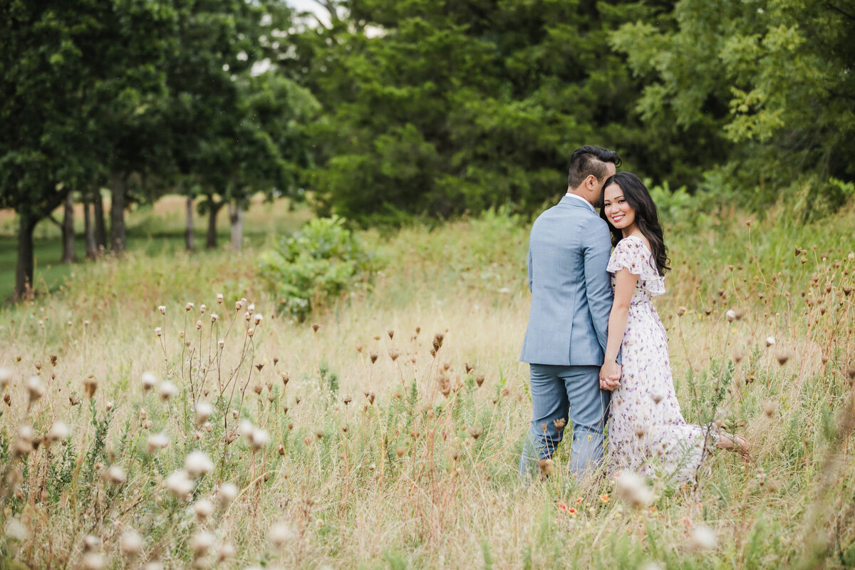 Couple standing in a field of wildflowers at White Rock Lake during their engagement session, she leans on her fiancé and looks back at the camera flirtatiously.