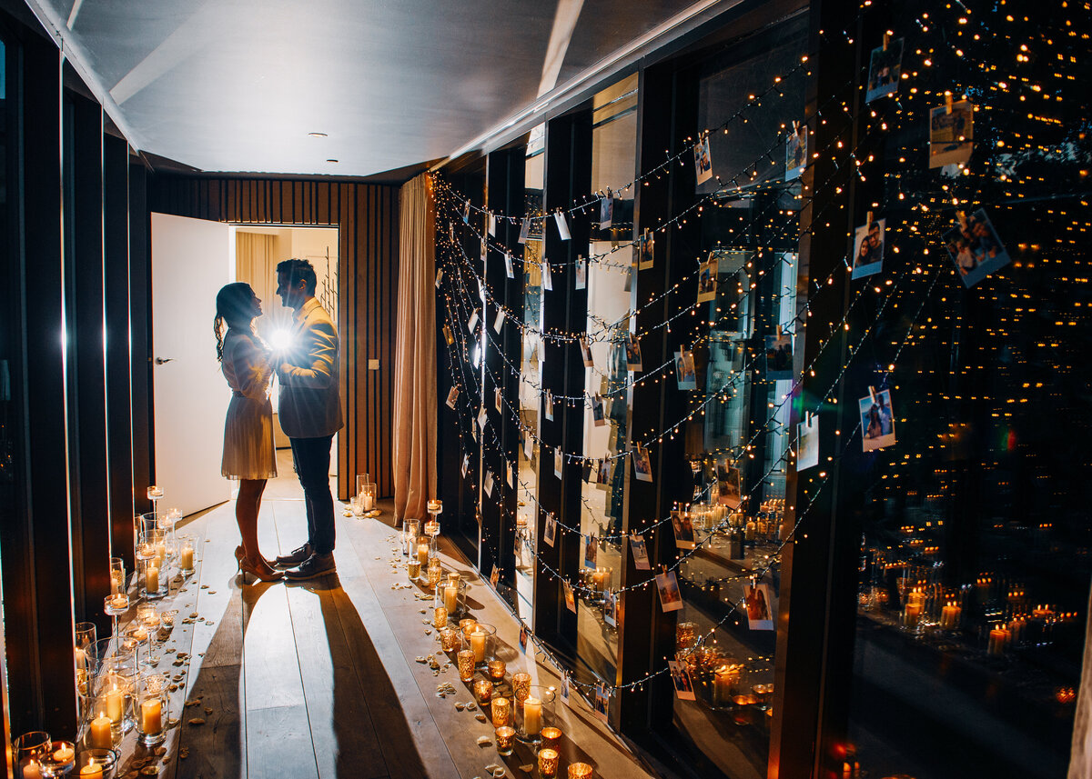 A couple stand holding hands in a hallway with fairy lights and polaroids with candles for a luxury wedding proposal.