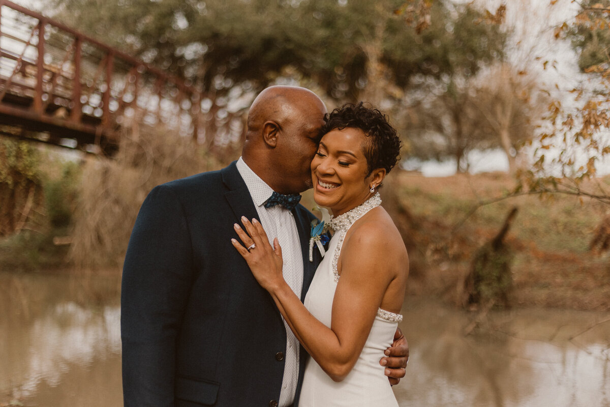Elopement portrait of a Black couple embracing at Oyster Creek Park, Sugar Land, Texas. The groom, wearing a dark suit, gently kisses the bride’s forehead while she smiles, dressed in a white gown. Captured during a romantic elopement by ShutterUp Photography.