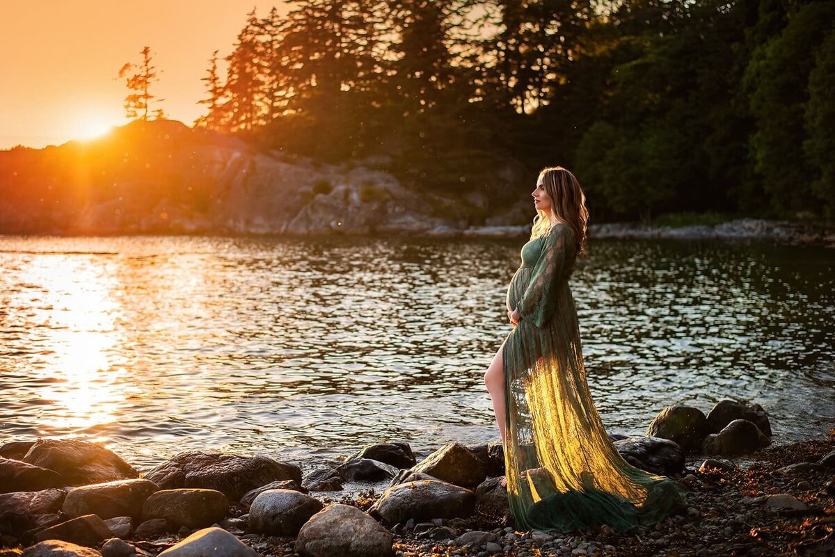 Pregnant woman in a green dress standing by the ocean at sunset during an outdoor maternity photoshoot at Whytecliff Park in West Vancouver.