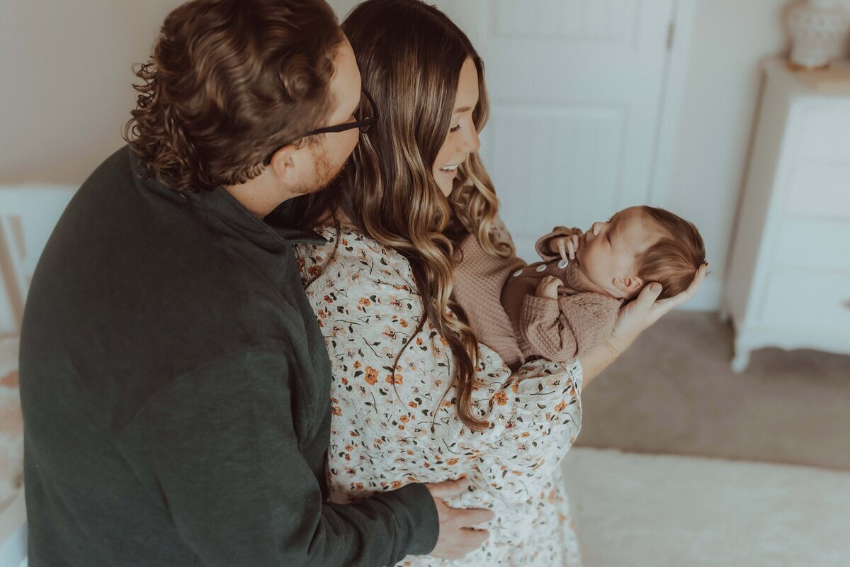 A man and woman smile at a baby they are holding. The woman cradles the newborn, while the man embraces her from behind. They stand in a softly lit room.