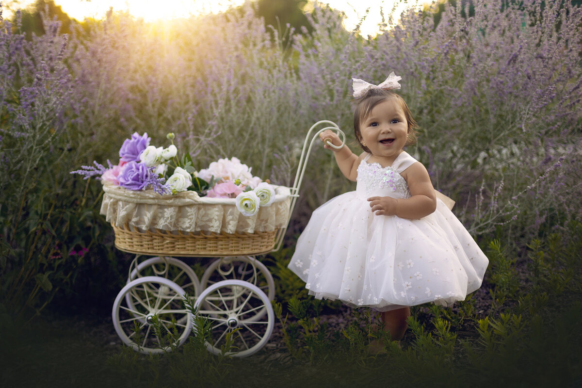 A young toddler girl in a white dress and matching bow pushes a tiny carriage full of flowers through a park at sunset