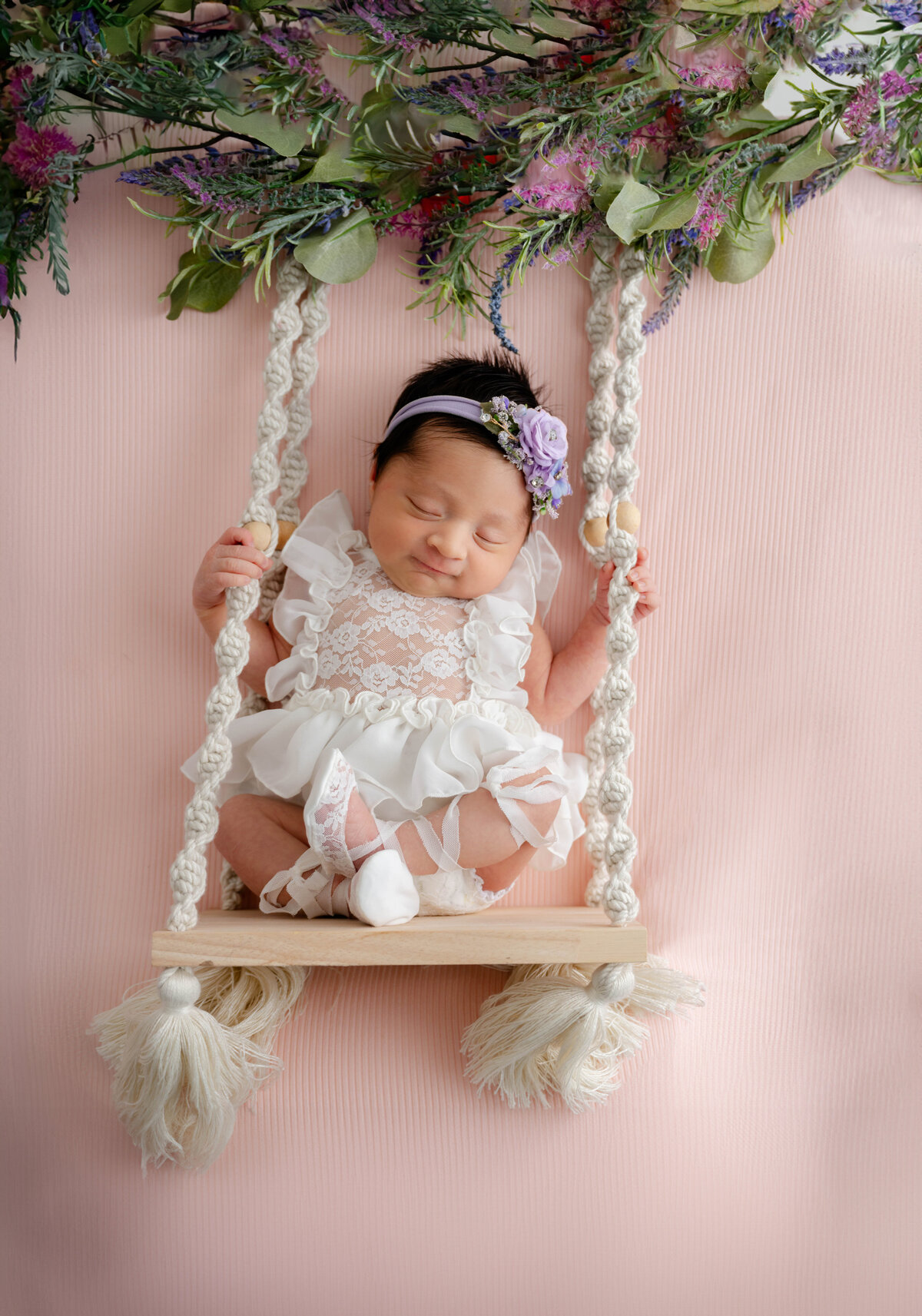 Smiling baby laying on a light pink backdrop, inside a swing, with her legs crossed.