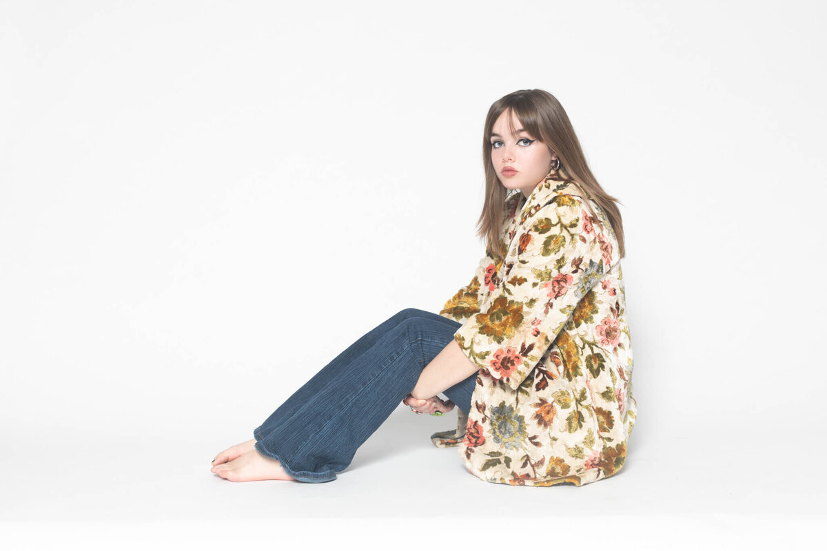 A young woman sits on the floor against a plain white background. She is wearing a floral-patterned top and dark blue jeans, with her legs bent and crossed. Her expression is neutral, and she gazes slightly to the side.