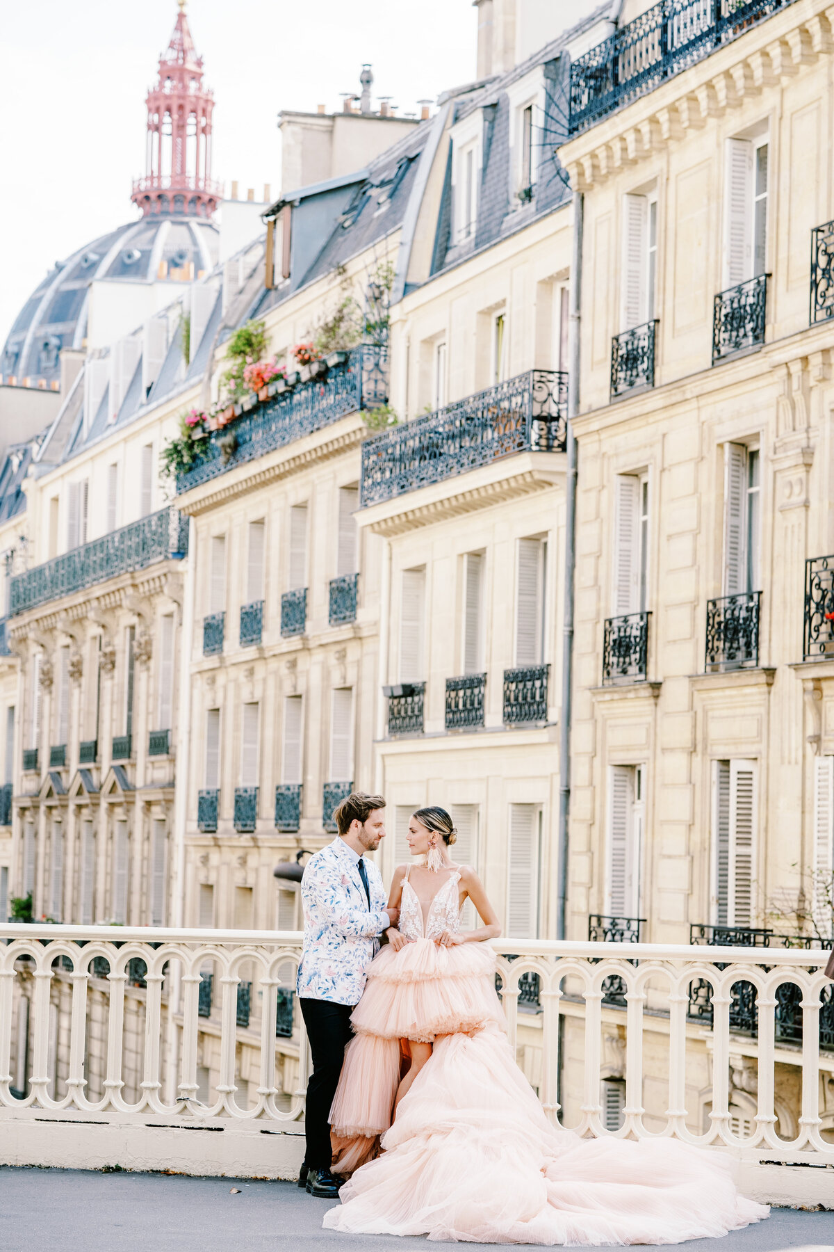 Couple on bridge with Paris homes behind them she is wearing a hi-lo peach gown leaning on the bridge looking at her future groom during their Paris engagement session, photographed by Italy wedding photographer.
