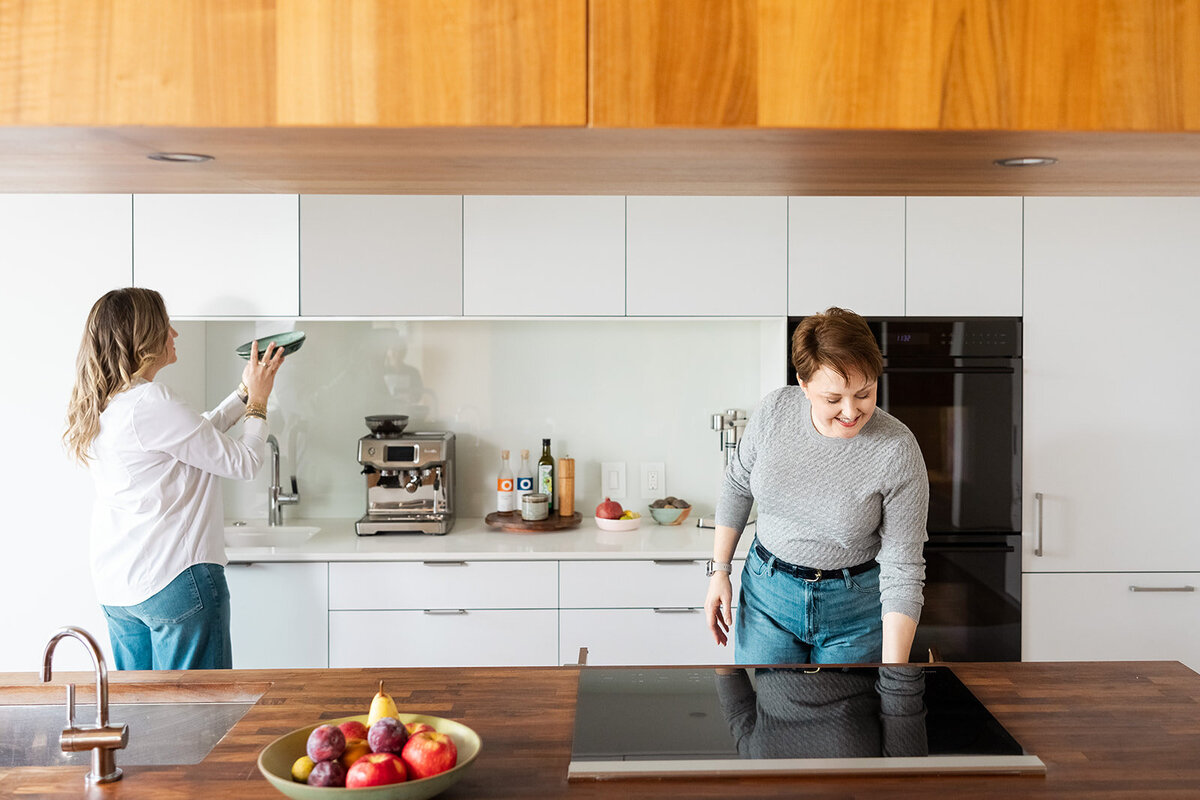 Two professional organizers wearing jeans and white tops organizing in a white kitchen.