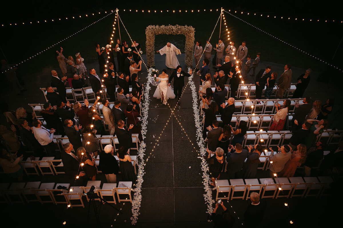 aerial photo of bride and groom walking down their ceremony aisle while everyone cheers in excitement photo by cait fletcher photography