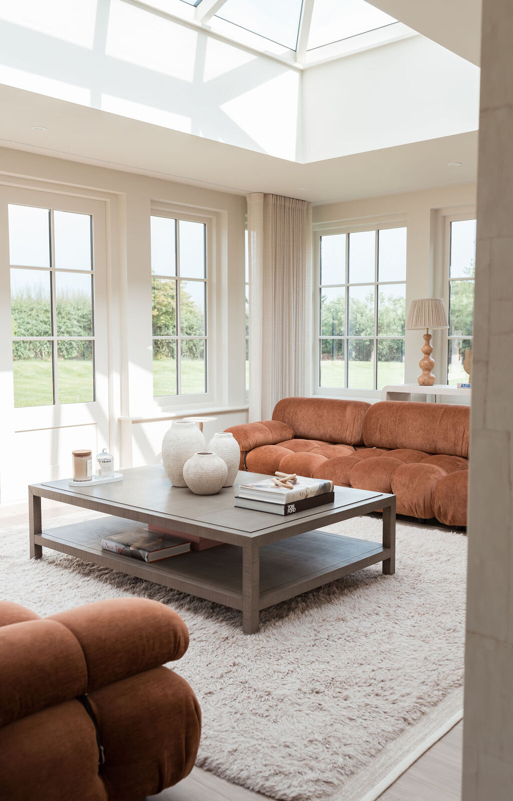 A cozy living room with large windows showcasing a greenery outside. The room features two rust-colored, plush sofas, a light grey coffee table with vases and books, and a soft, white rug covering the floor. Two lamps are on side tables flanking a large sofa.