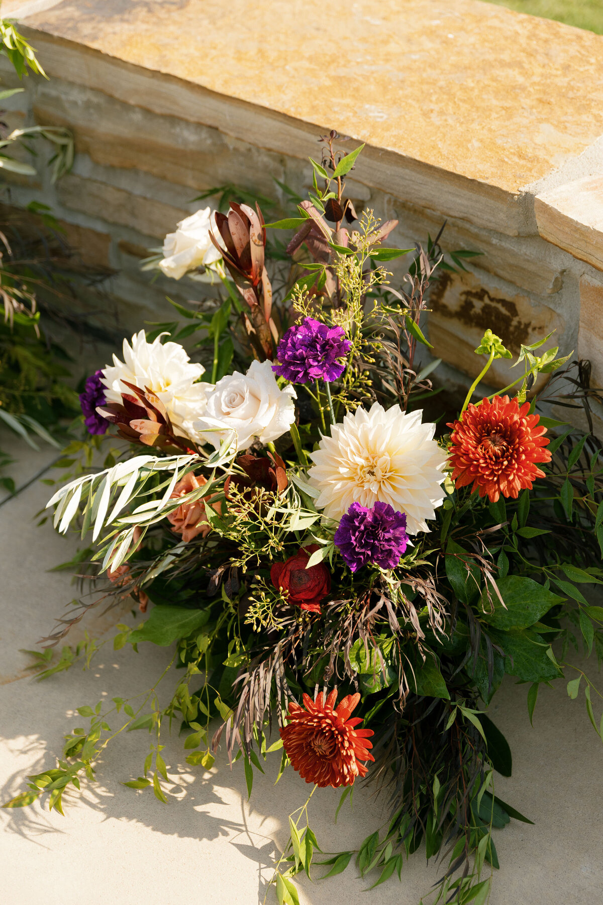 Lush outdoor wedding floral arrangement featuring ivory dahlias, peach roses, deep purple blooms, and greenery displayed against a stone ledge in warm sunlight at an elegant garden ceremony.