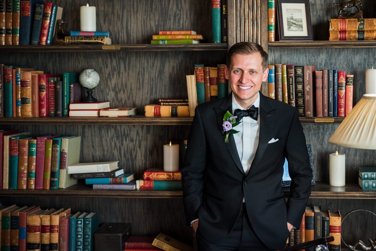 A groom leans up against the bookcase at the library in The Manor House in Littleton, Colorado.