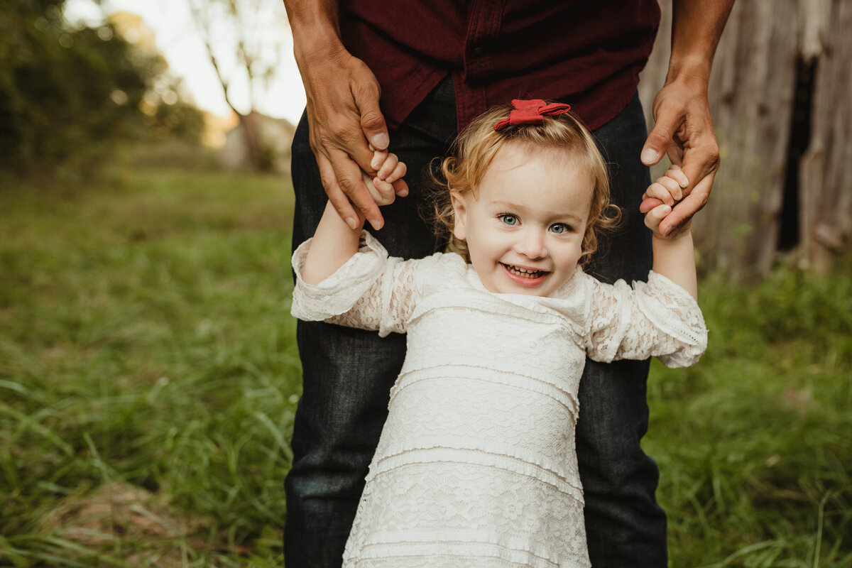 A little girl holds her dad's fingers while smiling.