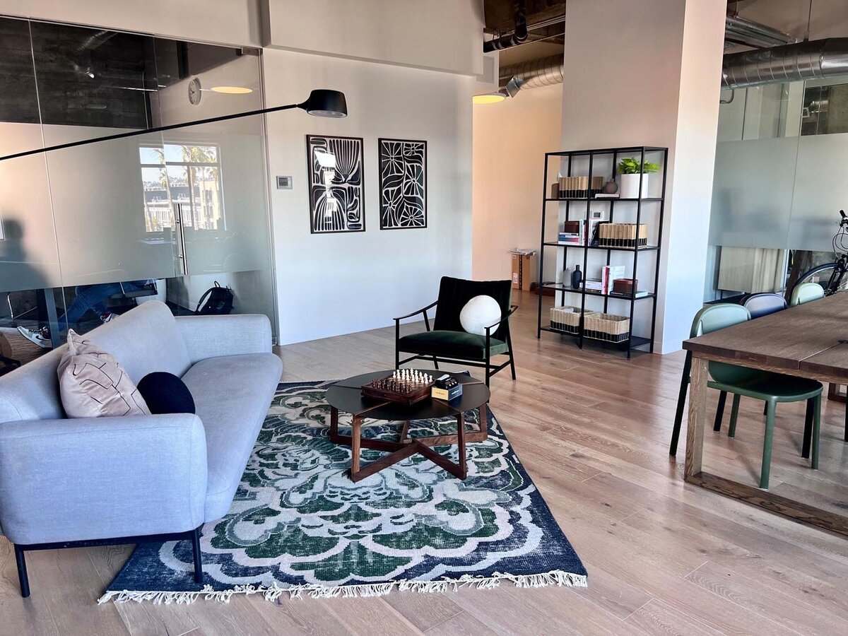 Open office lounge featuring a gray sofa, green velvet chair, patterned rug, black shelving unit, and wood dining table with green chairs.