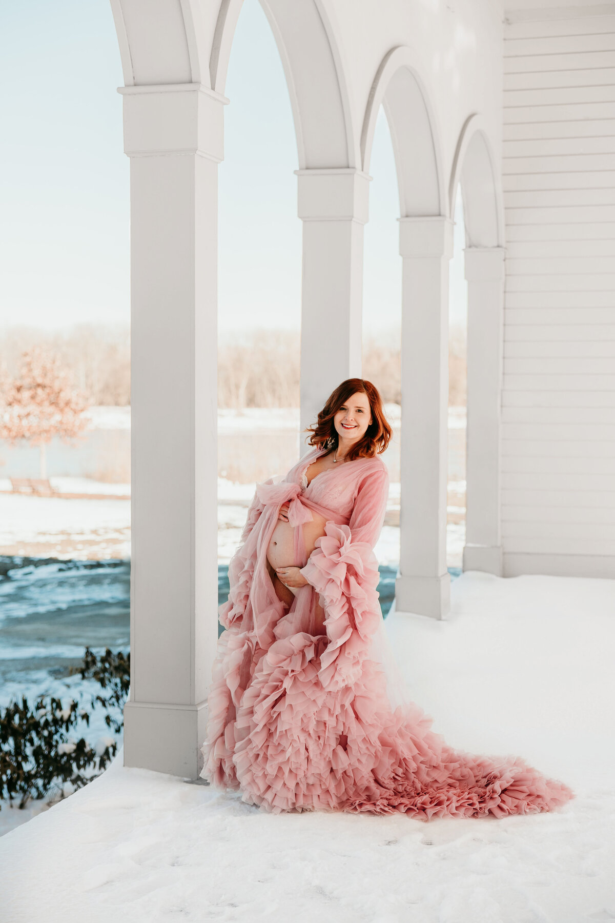 Pregnant mom stands in a snowy balcony wearing a flowy pink dress.