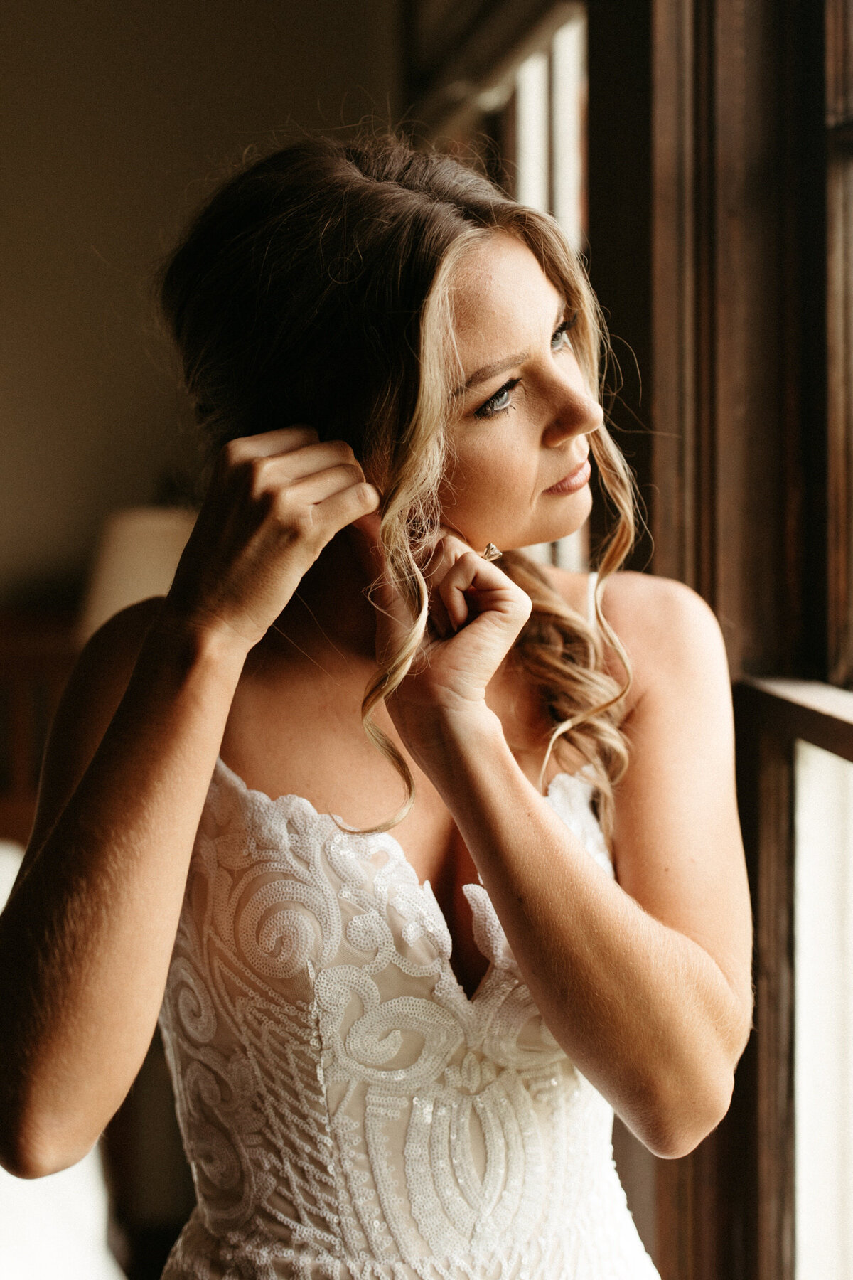Bride getting ready in front of a window putting her earring in on the morning of her wedding