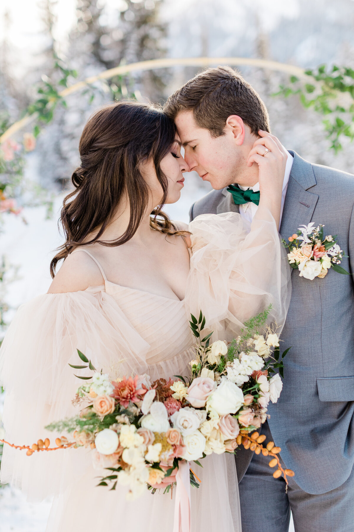 bride and groom stand together with eyes closed facing each other in peach wedding dress and grey suit with peach, orange, and white floral bouquet in Utah