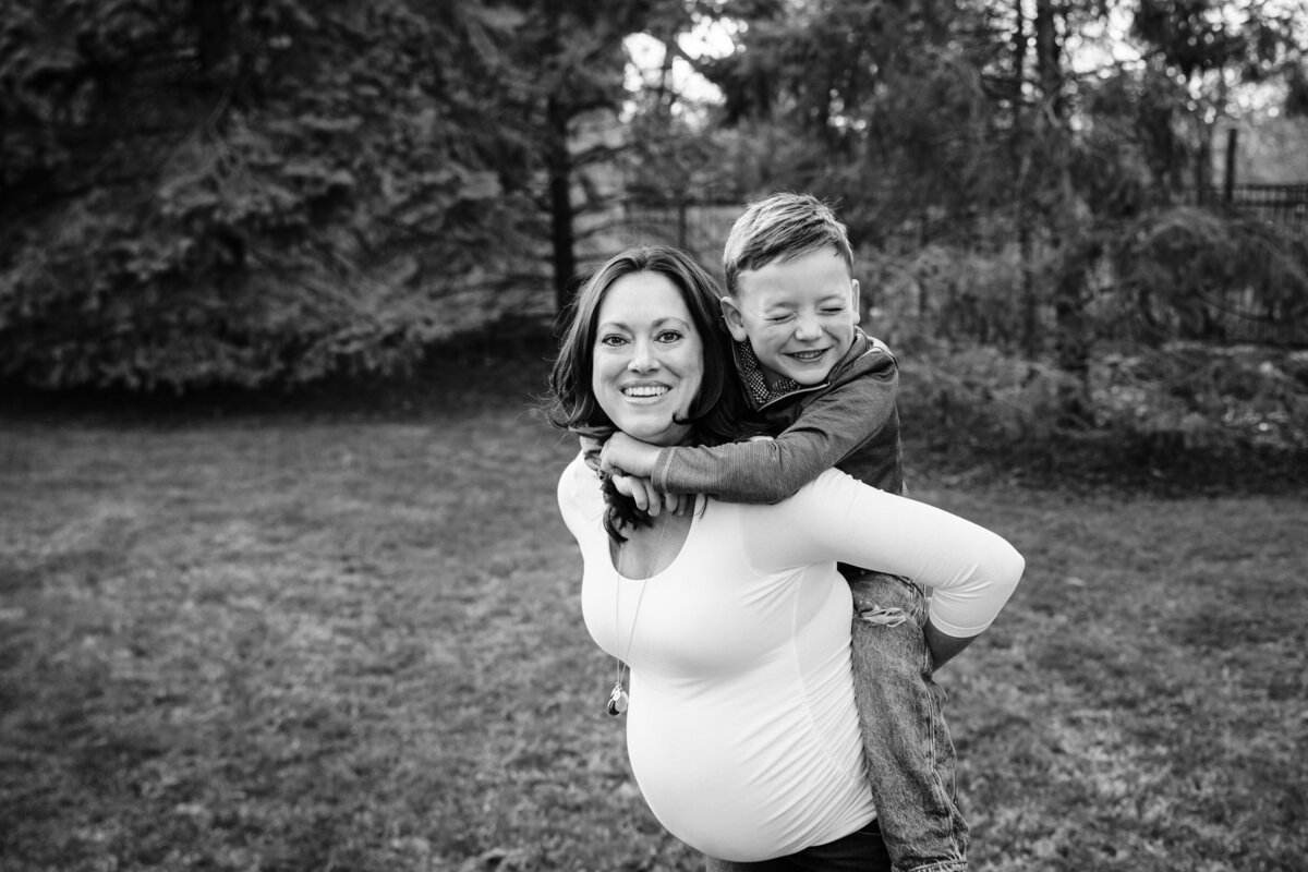 A pregnant woman, smiling and wearing a white top, carries her son on her back as they play in a grassy field.