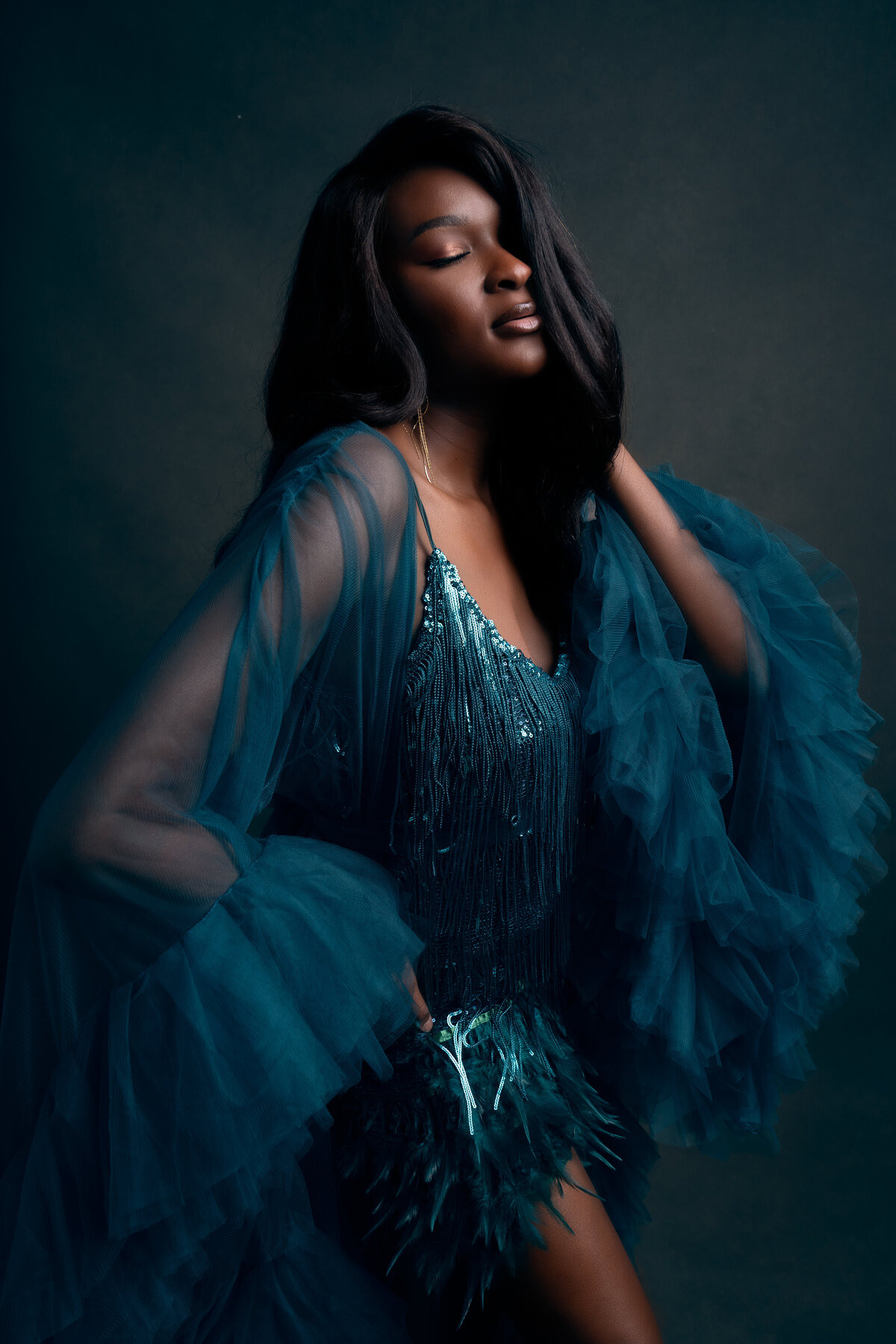 African American woman wearing blue-green sequin dress with tulle robe standing with eyes closed and left hand touching side of hair, right hand holding waist.
