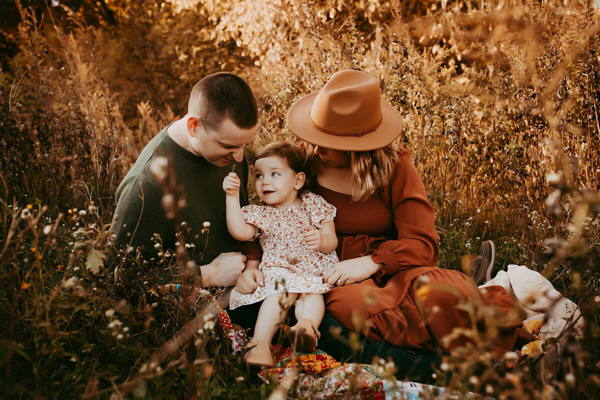 a little girl lifts a flower up to her dads nose so he can smell it in a field in rochester, ny