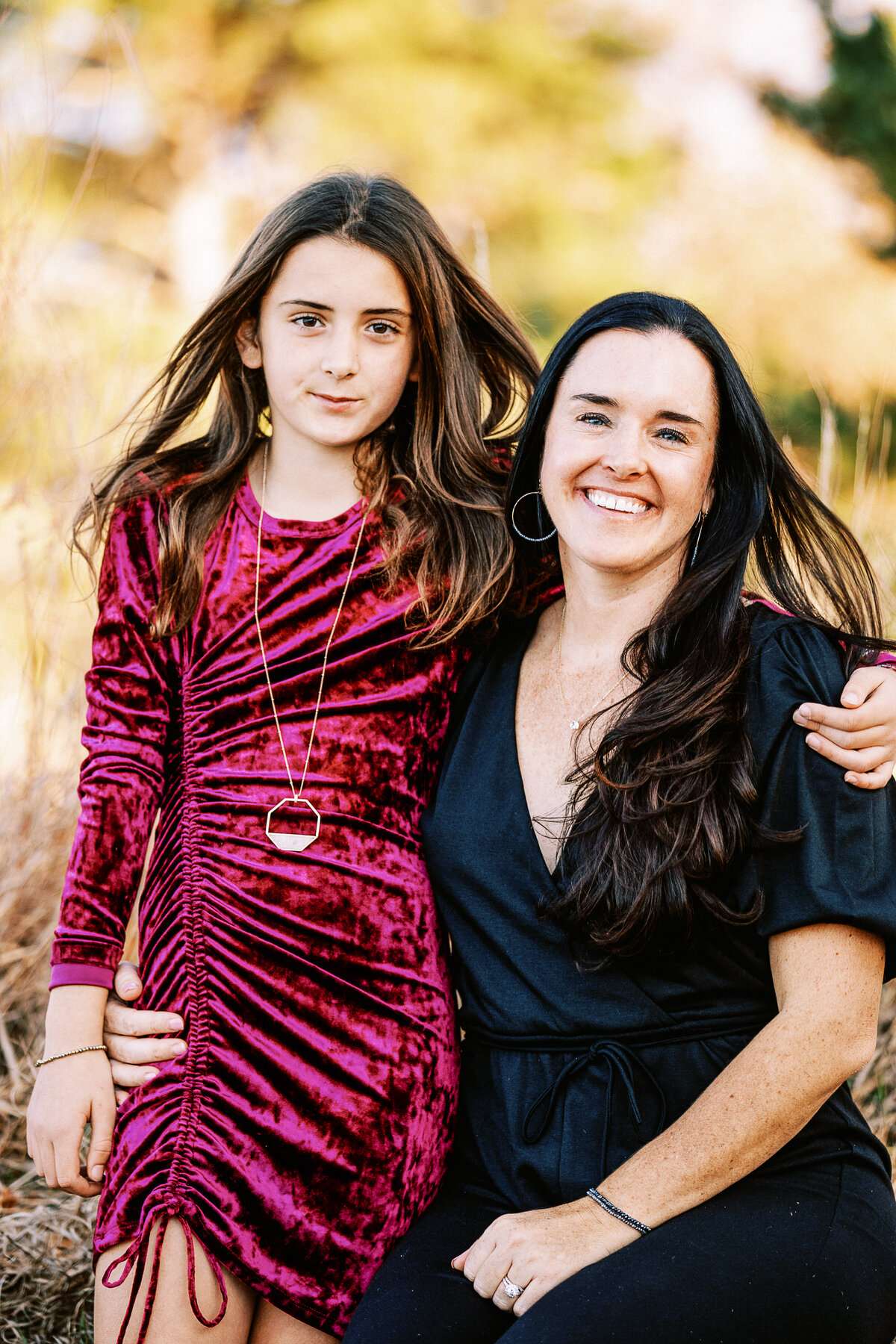 a girl in velvet dress stands next to her mom in a black jumpsuit for photos