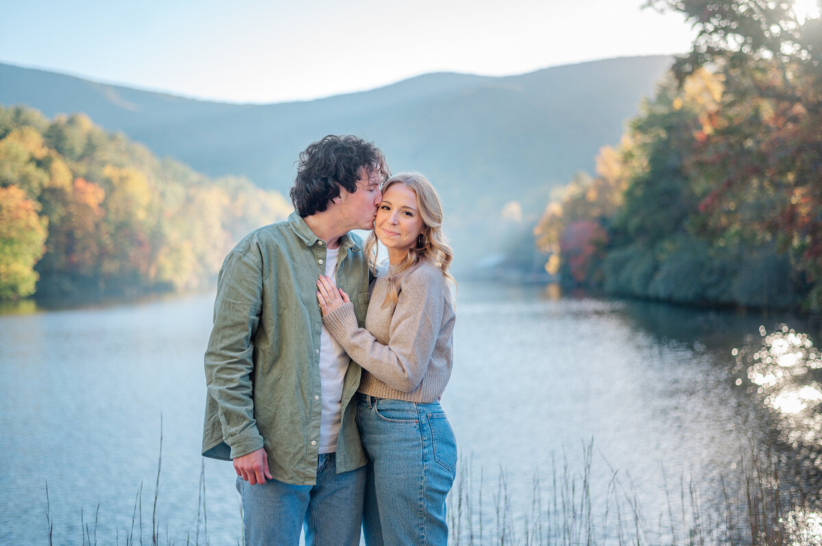 Man kisses his fiance on the cheek while she smiles. The beautiful North Georgia Mountains are in the background past the lake at Vogel State Park in Blairsville, Georgia