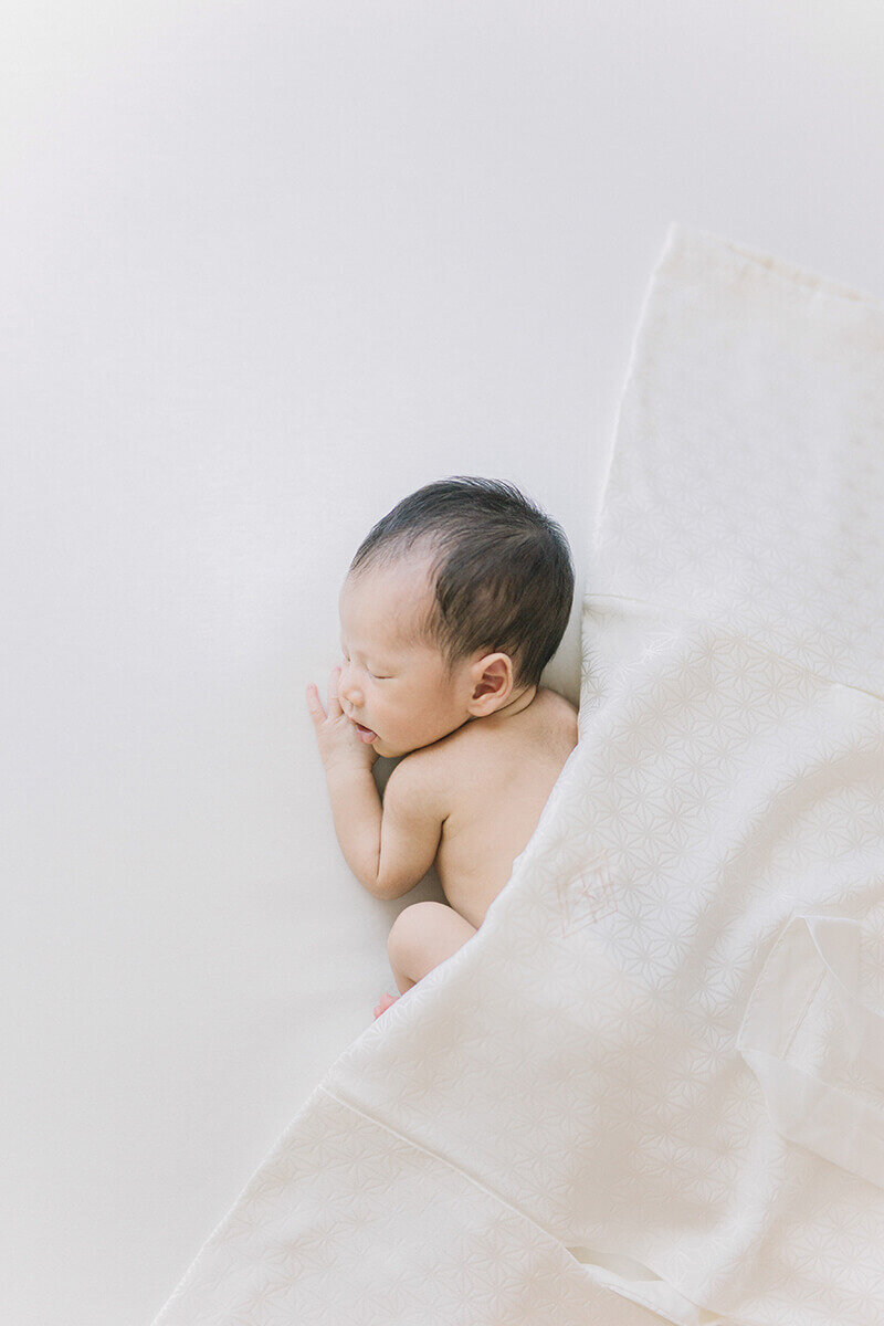 Newborn baby sleeping on his tummy with his mouth slightly opened. He has  pure white silk kimono over him and styled in a simple posed newborn shoot in Tokyo.