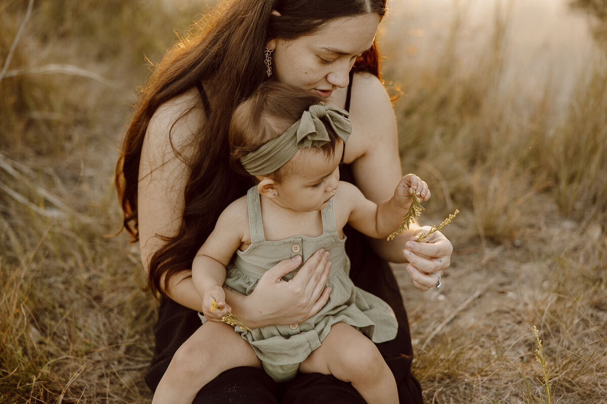 Mom and daughter sit in field and play with wildflowers