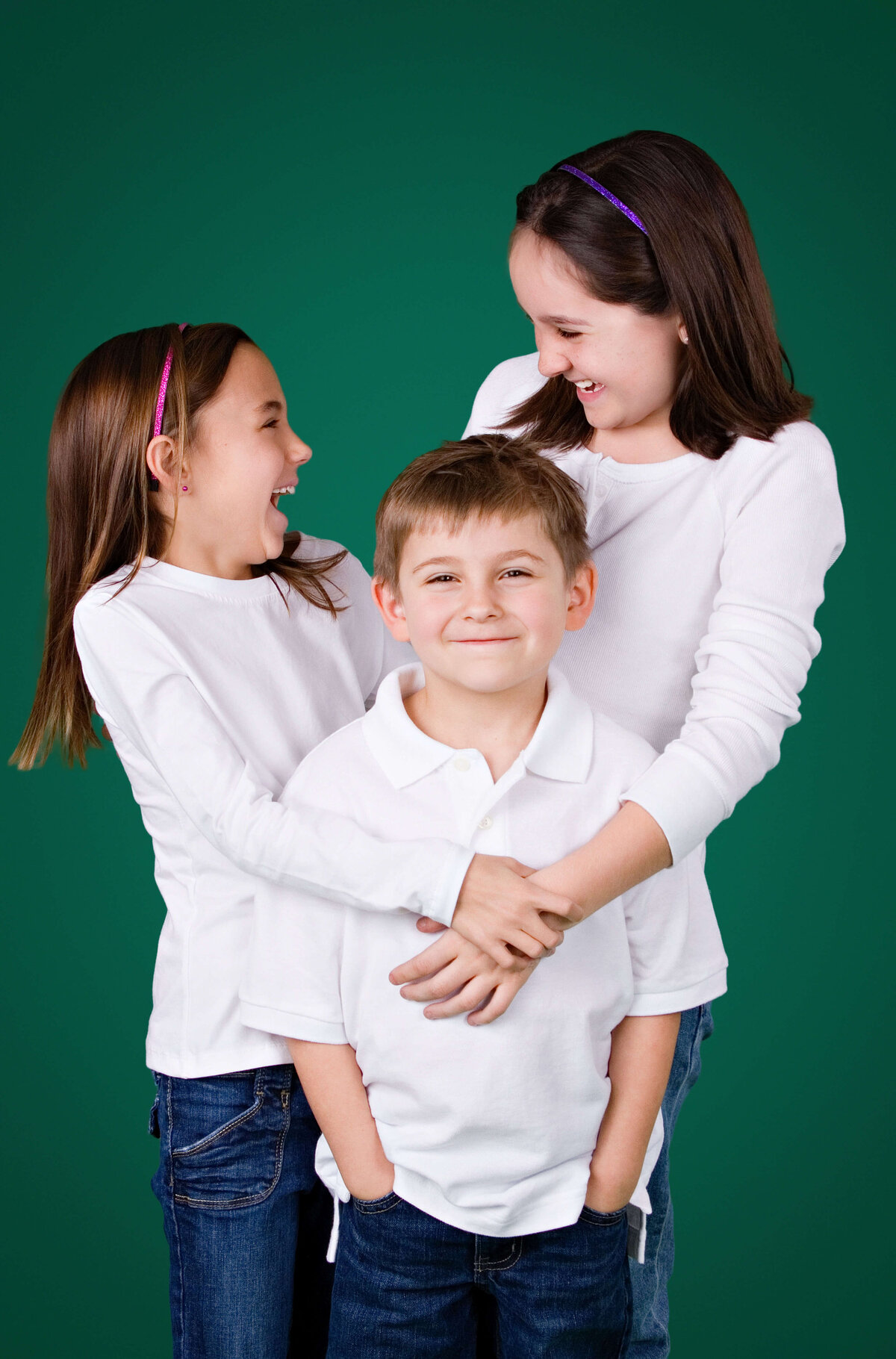 Three children dressed in white shirts and blue jeans stand against a green background. Two girls, wearing headbands, smile and laugh while standing beside a boy in the middle, who smiles with his hands in his pockets. Captured perfectly by Jacksonville family photography experts at the local portrait studio.