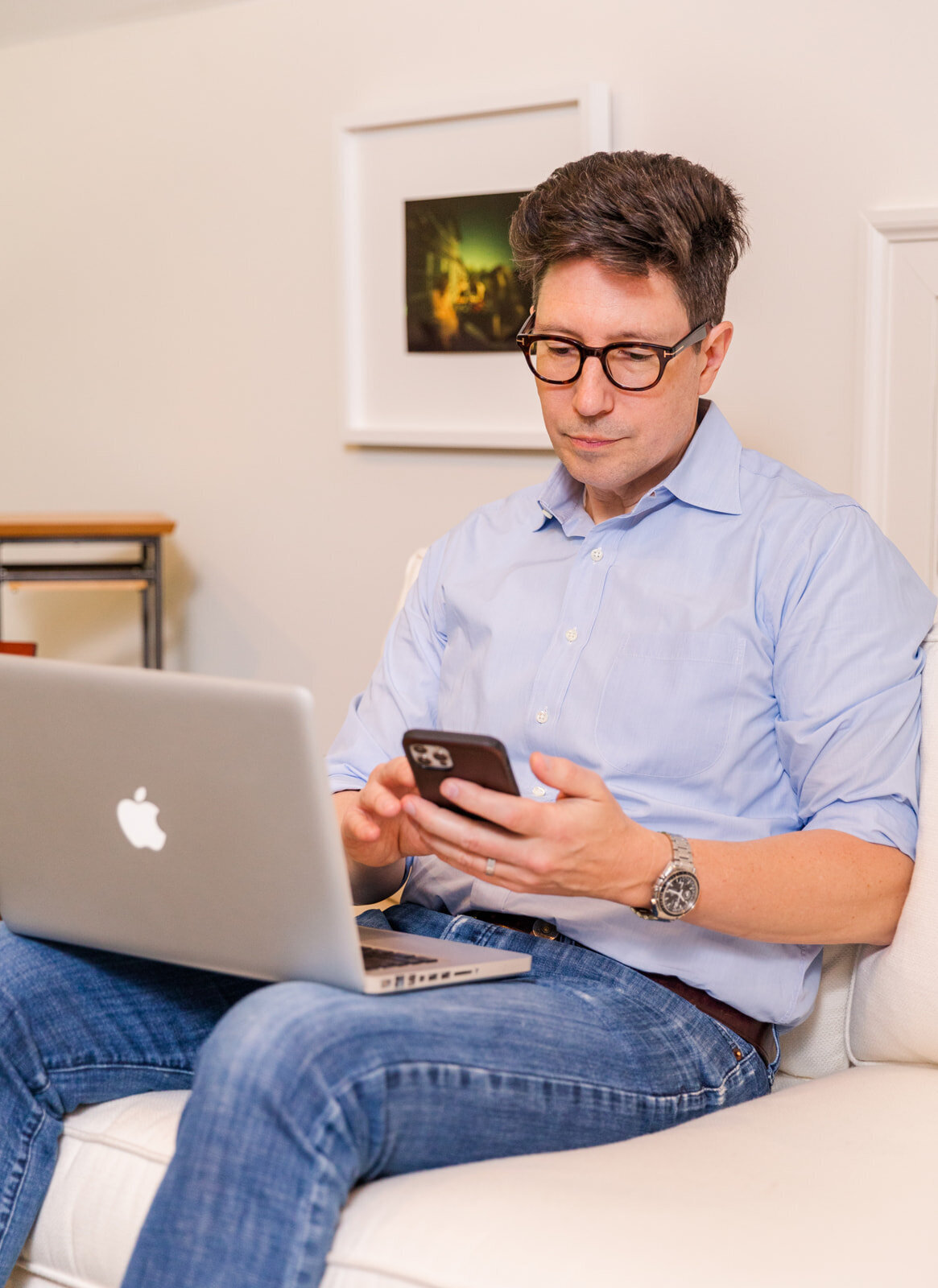 man in casual jeans and blue shirt sitting on a sofa working with his phone and laptop