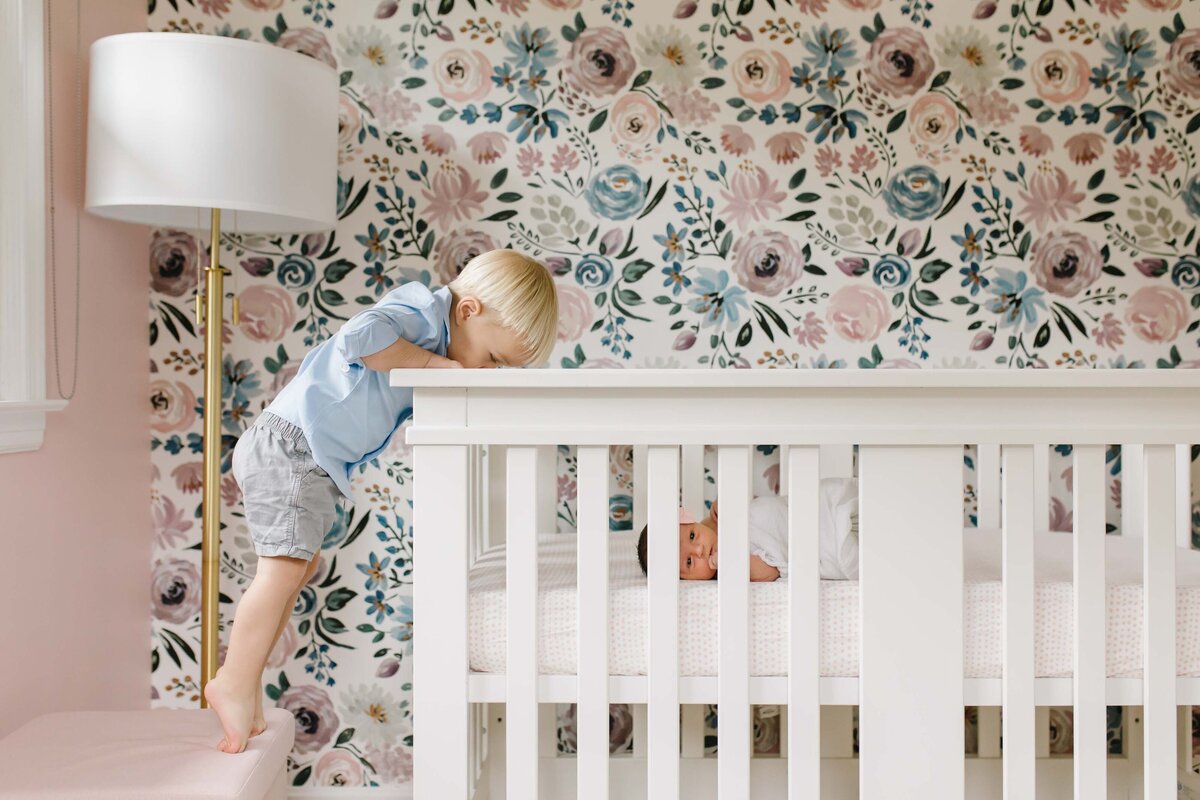 toddler boy standing on tippy toes looking into crib of newborn sister
